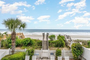 A wooden staircase leading to a beach area with a white picket fence, palm trees, and a wooden deck. - Beach Cabana: Oceanfront + Private Pool (Surfside Beach)