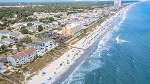 Une vue aérienne d’une ville côtière avec une longue plage bordée de parasols et de personnes se baignant, à côté d’une rangée d’immeubles à plusieurs étages dotés de balcons.