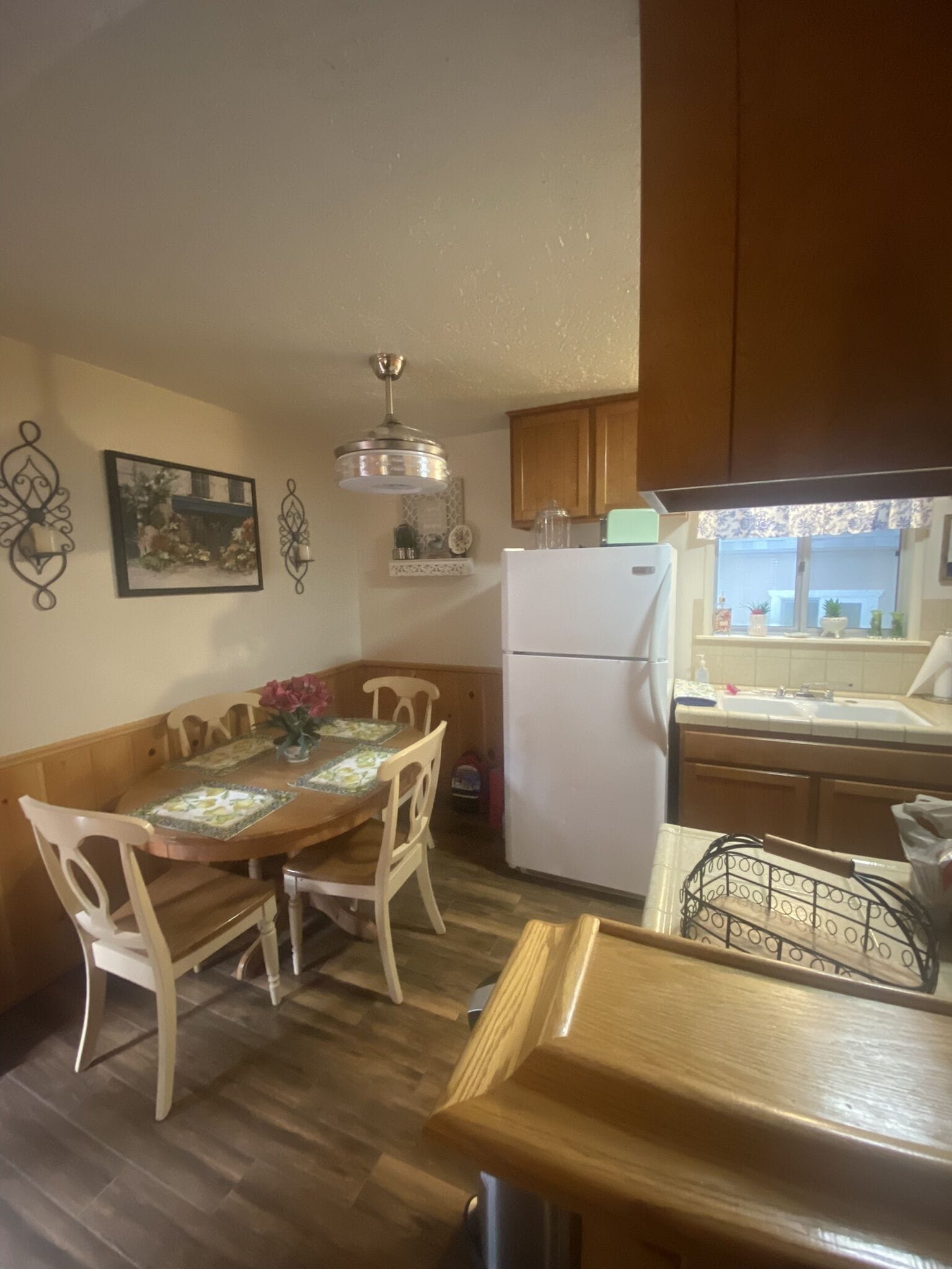 A compact kitchen with wooden cabinets, a white refrigerator, a dining table with chairs, and a hanging light fixture.