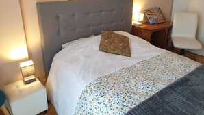 A neatly made bed with a grey headboard, a floral-patterned bedspread, and a decorative pillow. There is a bedside table with a lamp and a framed picture. A white chair is positioned next to the bed. - Spacious apartment - Paris St Denis, stade de France (Saint-Denis)