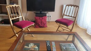 A living room with a flat-screen TV, two wooden chairs with red cushions, a glass coffee table with books and remote controls, and a bookshelf in the background. - Spacious apartment - Paris St Denis, stade de France (Saint-Denis)