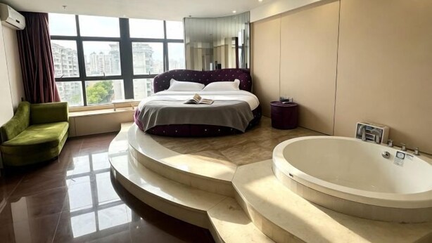 Une chambre moderne avec un grand lit, un canapé vert, une baignoire et une vue sur les immeubles à travers de grandes fenêtres.