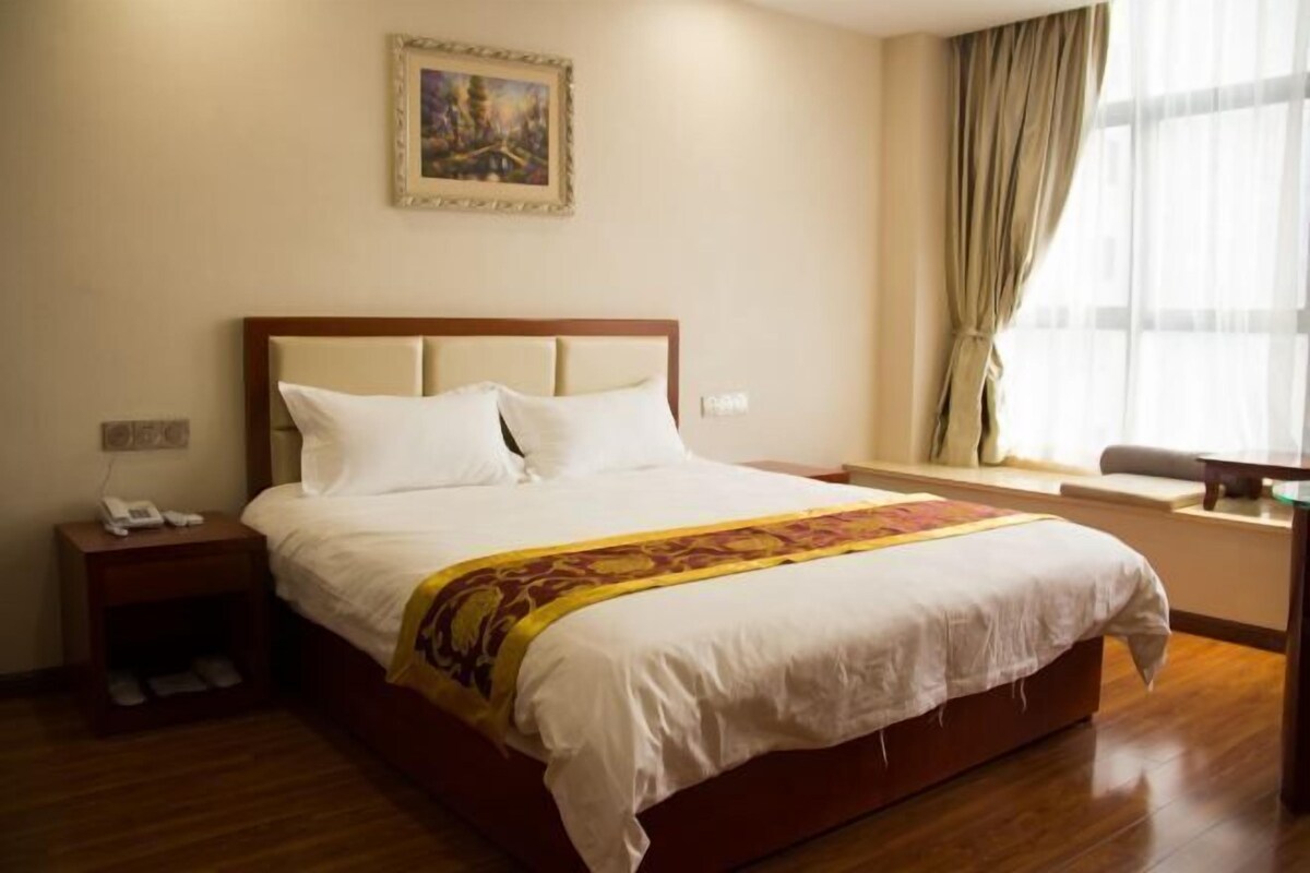 A neatly made bed with a white comforter and a decorative throw, a wooden nightstand with a phone, a painting on the wall, and a window with curtains.