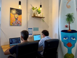 Deux personnes travaillent à des bureaux équipés d’ordinateurs dans une pièce où l’on aperçoit un tableau au mur, un panneau indiquant un espace de coworking et une étagère avec des plantes.