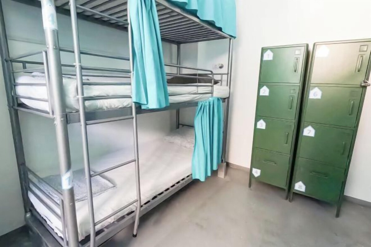Una cama individual con cortina azul, un casillero verde con etiqueta numérica y suelo de hormigón.