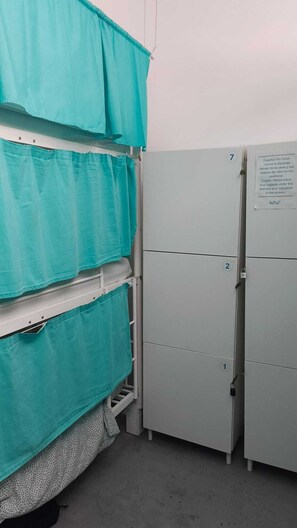 Una habitación con una cama cubierta con sábanas color turquesa, un armario metálico con cerradura y un aviso en la puerta.