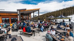 Un espacio al aire libre con mesas y bancos, una zona cubierta y el paisaje de montañas al fondo.