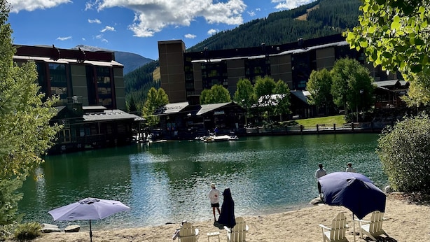 Vista de un lago con un conjunto de edificios, sillas y una sombrilla en la arena.