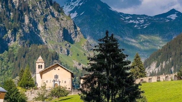 Une église de montagne dotée d’un clocher, entourée d’une végétation luxuriante et de falaises abruptes.