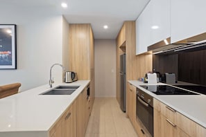 Cocina moderna con encimeras blancas, armarios de madera y electrodomésticos de acero inoxidable, incluyendo un horno empotrado.