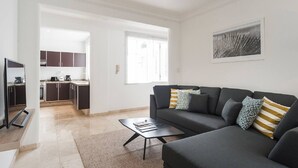 Una sala de estar moderna con un sofá gris oscuro, una mesa de centro y un televisor. Cuenta con una zona de cocina con armarios oscuros y un ventanal.