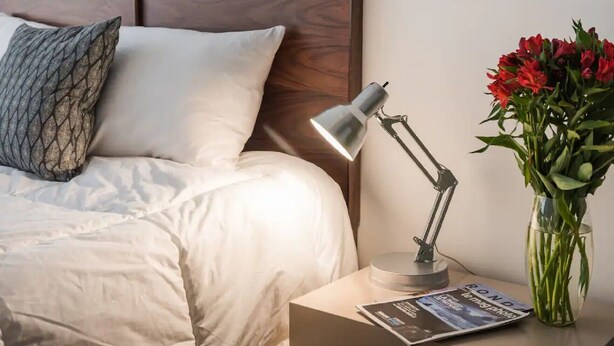 Una cama bien hecha con sábanas blancas, una almohada gris estampada, una lámpara de noche, un jarrón con flores rojas y una revista sobre una mesita de noche.