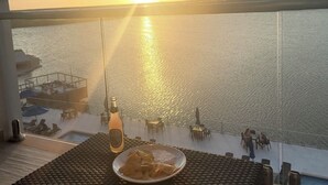 Un balcón con una mesa, una botella y un plato de comida, con vista a un cuerpo de agua durante el atardecer.