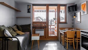 Ein kompakter Wohnbereich mit einer Couch, einem kleinen Tisch, einer Essecke mit Stühlen, einem an der Wand montierten Fernseher und einer Küchenzeile mit Backofen.