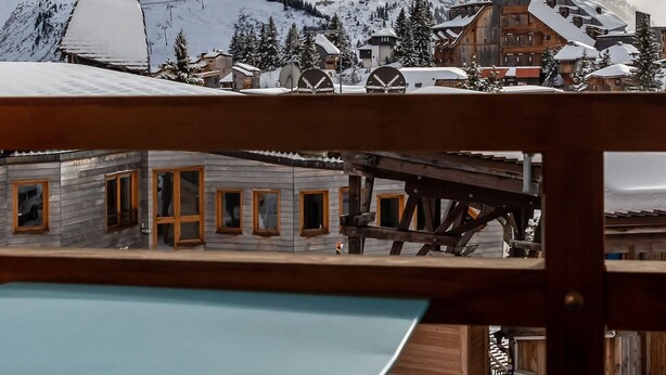 Eine Holzterrasse mit Tisch, mit Blick auf eine gebirgige Landschaft mit schneebedeckten Gebäuden und einer Seilbahn.