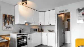Eine moderne Küche mit weißen Schränken, einem eingebauten Backofen und einer Mikrowelle. Ein Esstisch mit Stühlen ist vorhanden, und ein gelber Stuhl ist im Vordergrund sichtbar.