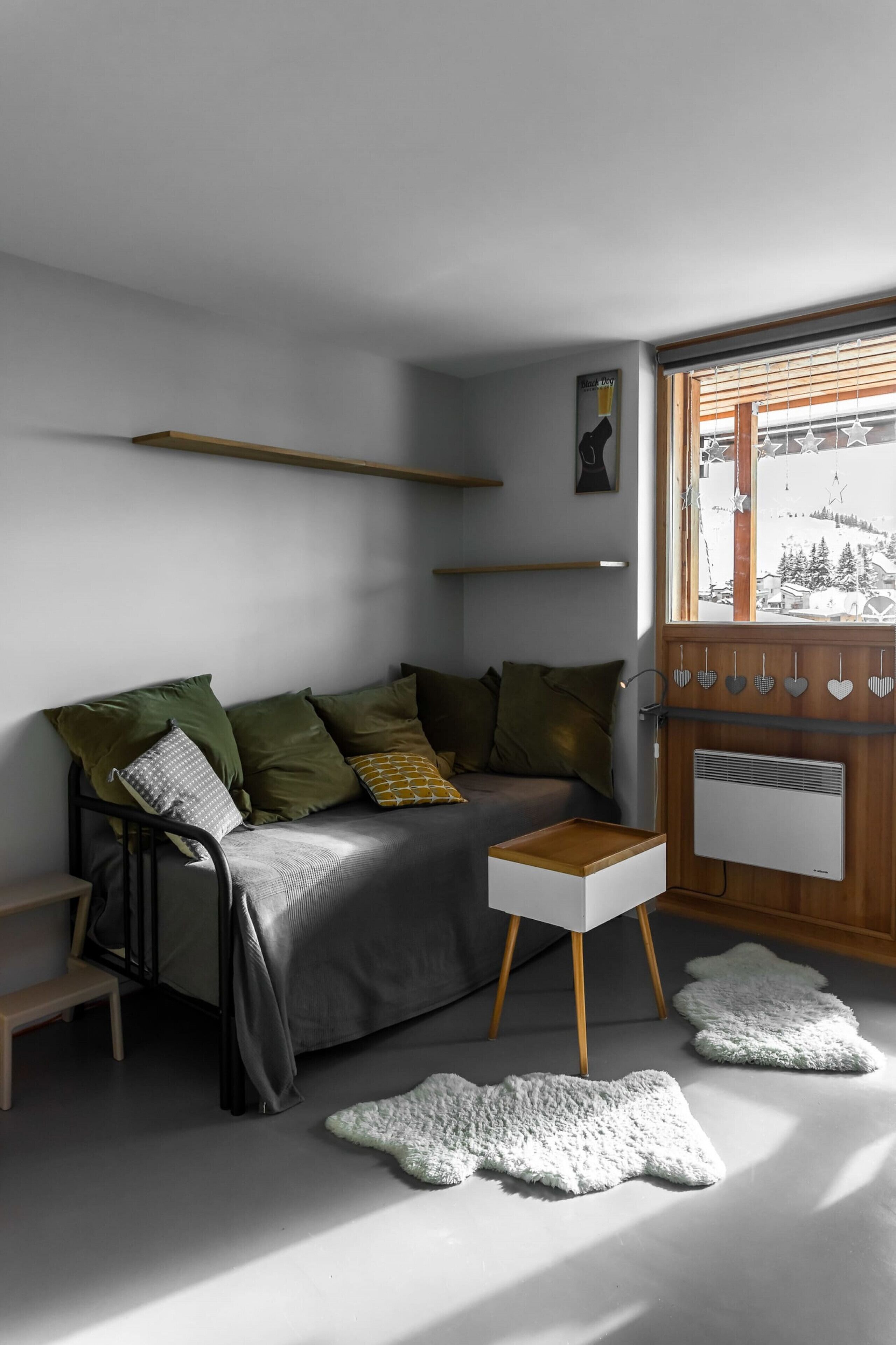 Ein modernes Wohnzimmer mit grauem Sofa, Holznachttisch und einem Fenster mit sternförmigen Jalousien.