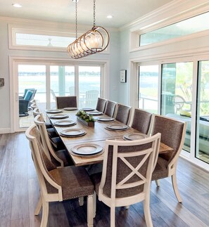 Enjoy meals in this dining area with a long table, high ceiling, and large windows offering a view of the outdoors.