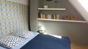 A bedroom with a bed, patterned pillows, a nightstand with a lamp, and bookshelves with books and baskets. - Charming cottage "La Casa Céline", near the sea (Surtainville)