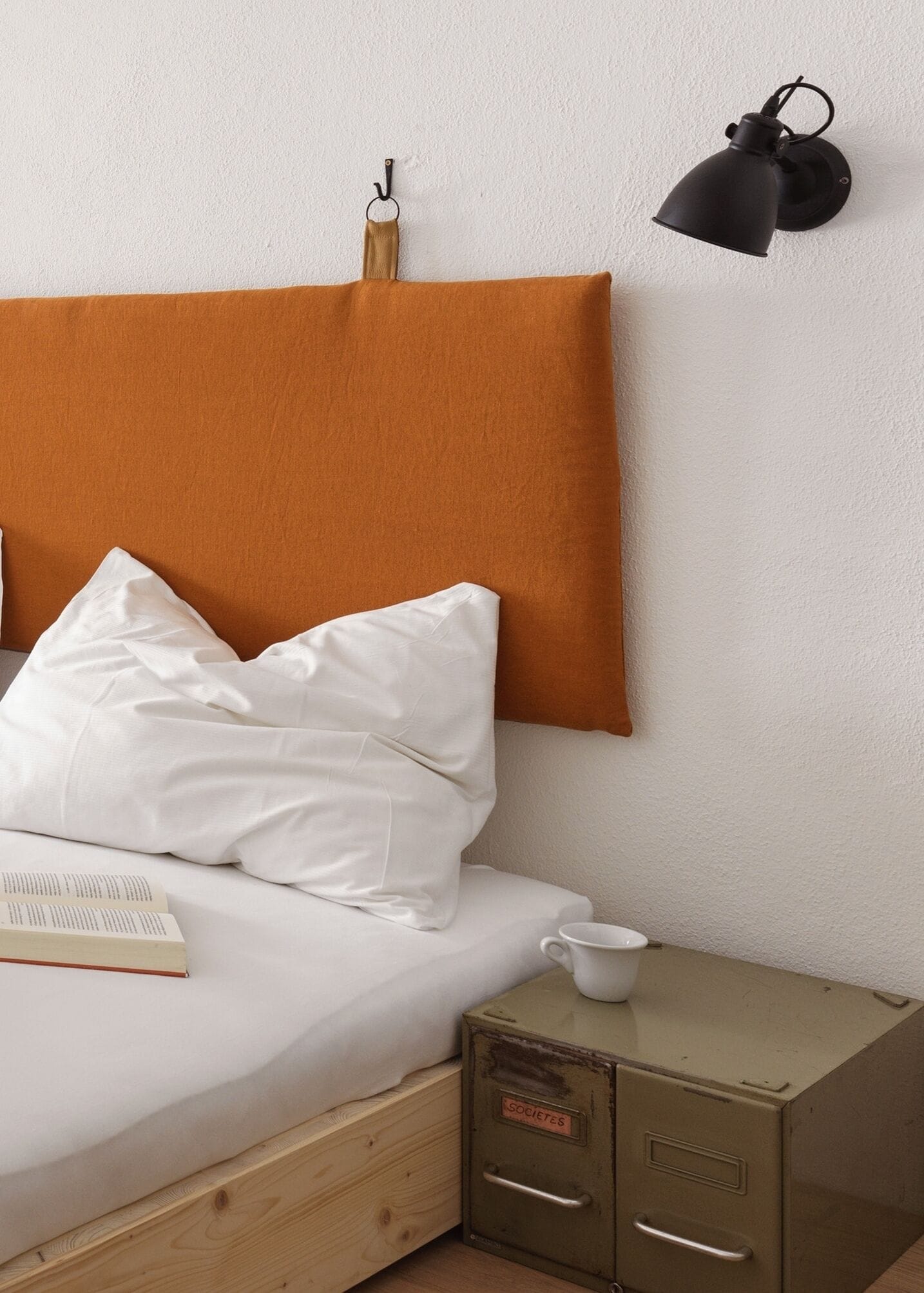 Un letto con biancheria da letto bianca, una testiera marrone, una lampada da parete nera, una tazza bianca e un comodino in legno con una cassettiera metallica.
