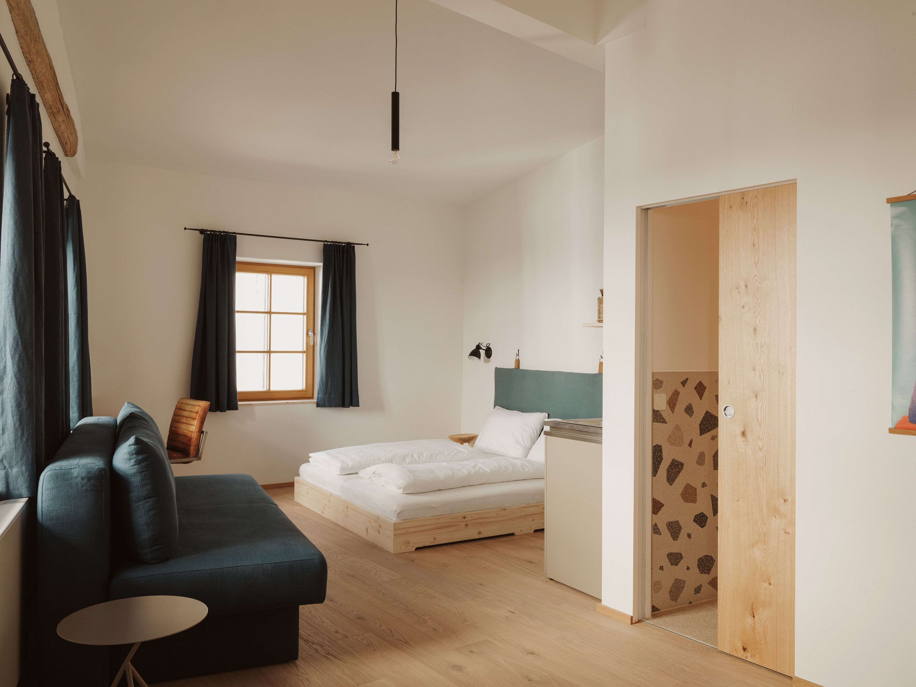 A modern bedroom with a wooden floor, a bed with white bedding, a dark blue sofa, and a small round table.