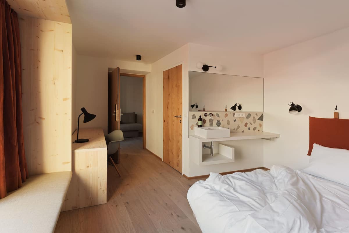 A hotel room with a wooden floor, a bed, a desk with a lamp, a bathroom with a sink and mirror, and a door leading to a balcony.