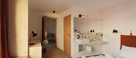 Una camera d'albergo con pavimento in legno, un letto, una scrivania con lampada, un bagno con lavandino e specchio, e una porta che dà su un balcone.