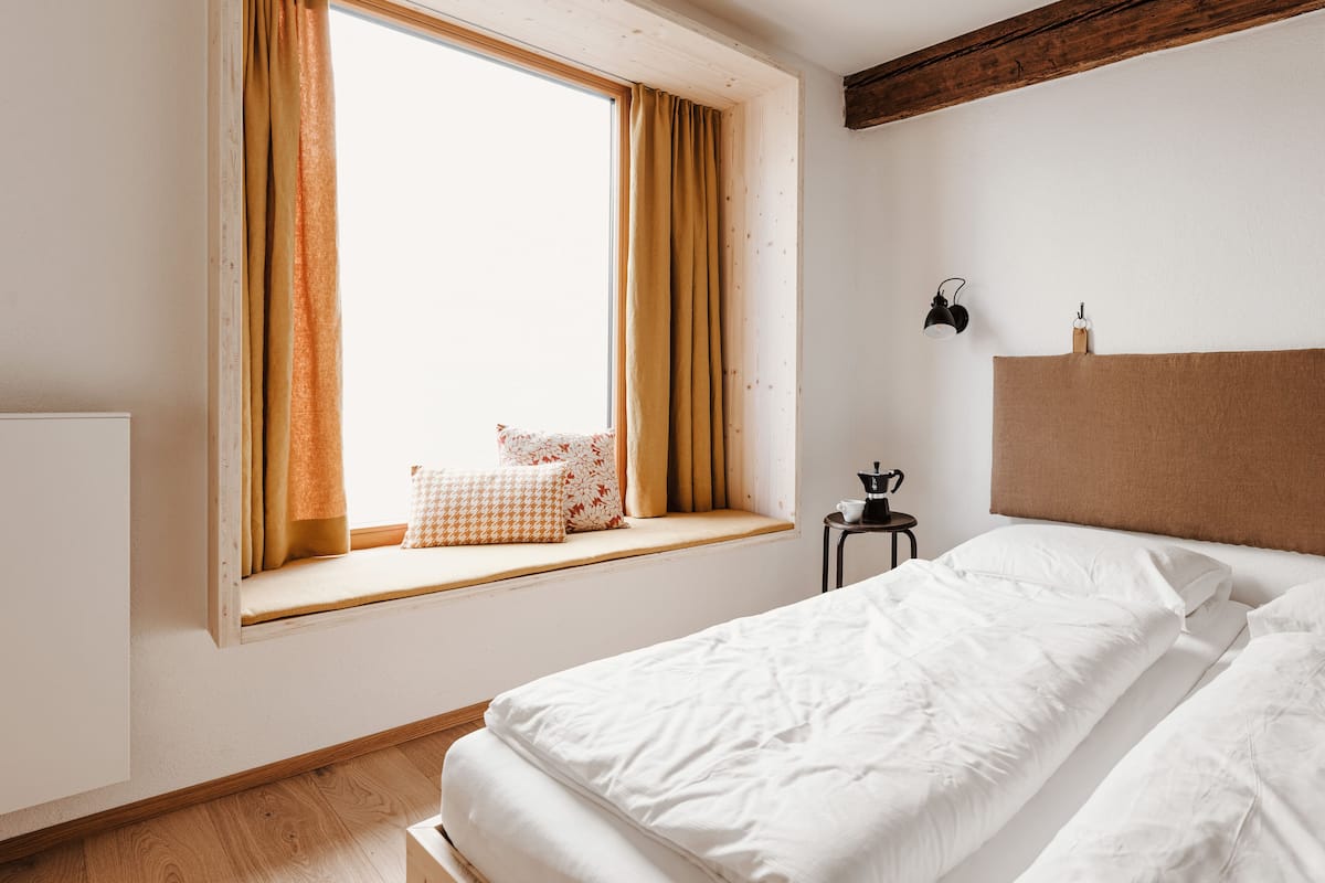 A bedroom with a large window, a bed with white linens, a small bedside table with a coffee pot, and a headboard.
