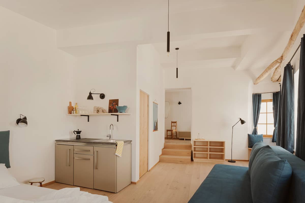 A modern, well-lit room with a kitchenette, a bed, a sofa, and a view of a cityscape through the window.