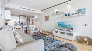 A modern living room with a sofa, coffee table, and TV. There is a kitchen in the background. - Villa-style 2BR Condo w Pool, GYM Pass near Beach (Noord)