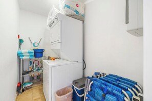 A laundry area with a washing machine, shelves with cleaning supplies, and a trash bin.