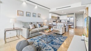 A modern living room with a grey sofa, coffee table, and a blue rug. There is a kitchen in the background with white cabinets and a dining area.
