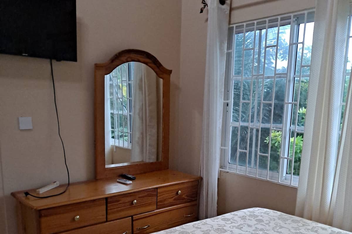 A bedroom with a wooden dresser, a bed with a patterned bedspread, a wall-mounted TV, and a window with a view of greenery.