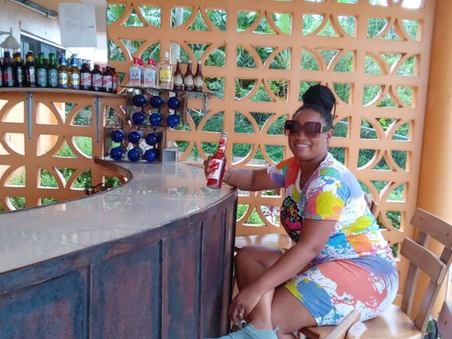 Una persona sentada en una barra con mostrador y sillas de madera. Hay botellas y vasos sobre el mostrador, y un panel de madera decorativo con un diseño geométrico al fondo.