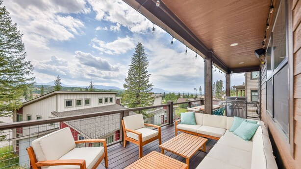Um terraço coberto ao ar livre com móveis de madeira, incluindo um sofá e cadeiras, com vista para uma área residencial com casas e árvores.