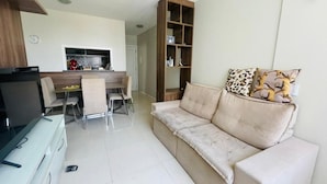 A living room with a sofa, a dining table with chairs, a kitchen area, and a bookshelf.