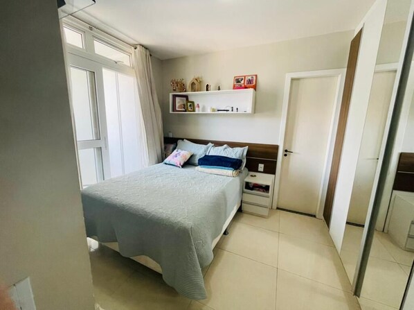 A bedroom with a bed, a nightstand, a shelf with personal items, and a door. - Excelente Apartamento_papicu (Fortaleza)