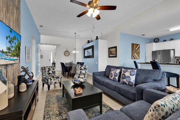 A modern living room with a sofa, coffee table, and decorative items. There is a dining area with chairs and a table, and a kitchen with cabinets and appliances.