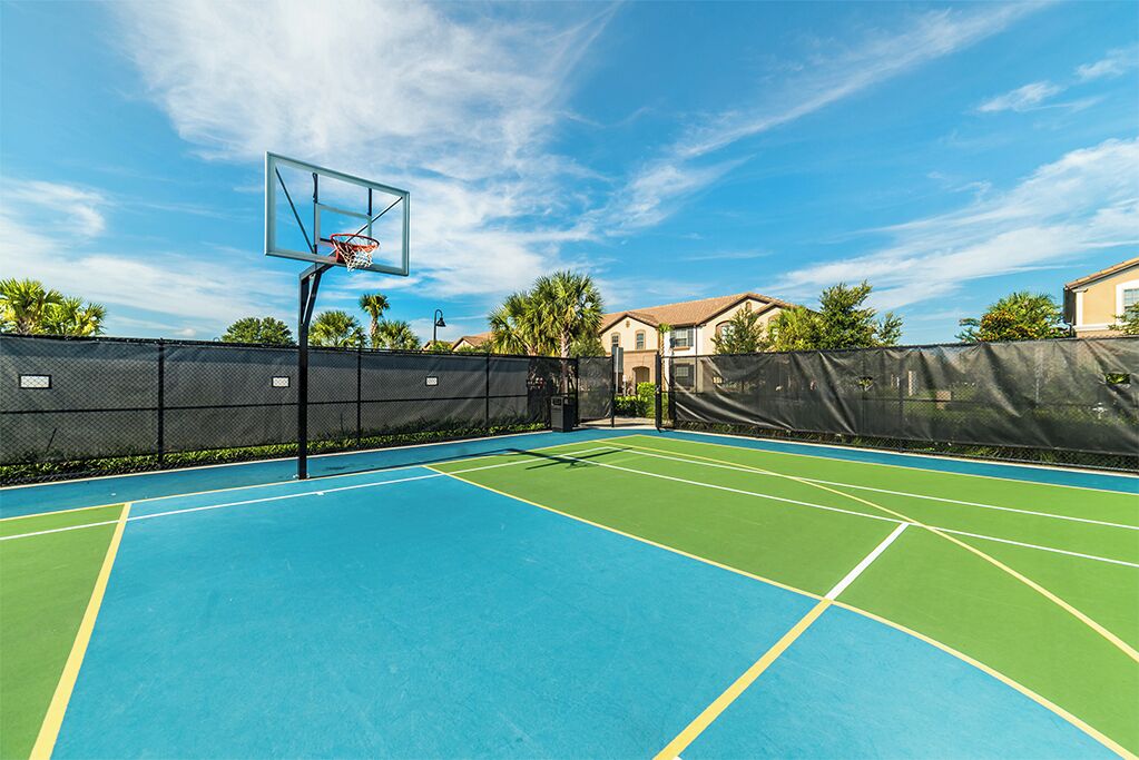 An outdoor basketball court with a net, surrounded by a fence, and houses in the background.