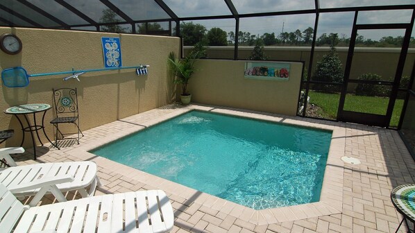 A swimming pool with lounge chairs and a small table, surrounded by a brick patio and a glass enclosure.