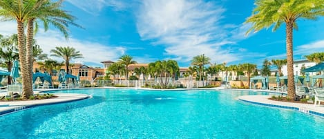 A large outdoor swimming pool surrounded by palm trees and lounge chairs, with a clear blue sky above.