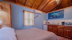 Un dormitorio con cama, techo de madera, ventana con cortinas, aire acondicionado, zona de cocina con microondas y un cuadro en la pared.