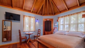 Un dormitorio con techo de madera, una cama con cubrecama acolchado, un televisor montado en la pared, un espejo, una mesita pequeña, dos sillas y una ventana con cortinas.