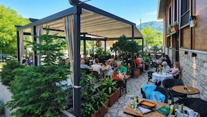 Een terras met tafels en stoelen, omgeven door groen en een overdekte zithoek.