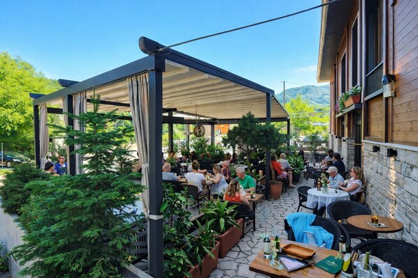 An outdoor patio area with tables and chairs, surrounded by greenery and a covered seating area.
