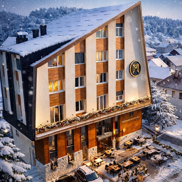 A modern hotel with a snow-covered roof, a restaurant on the ground floor, and a view of a snowy village in the background.