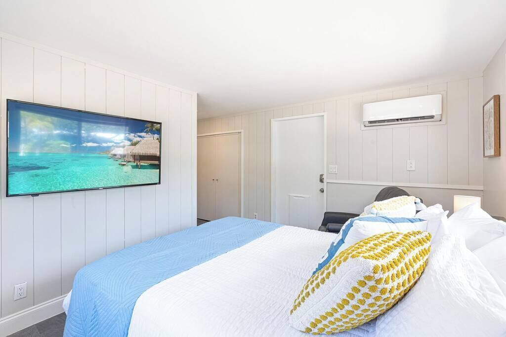 A hotel room with a bed, a television displaying a tropical beach scene, an air conditioning unit, and a door.