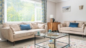 Sala de estar con dos sofás beige, una mesa de centro de vidrio, un tapiz estampado y una ventana con persianas.