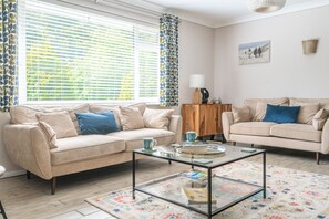 A living room with two beige sofas, a glass coffee table, a patterned rug, and a window with blinds.