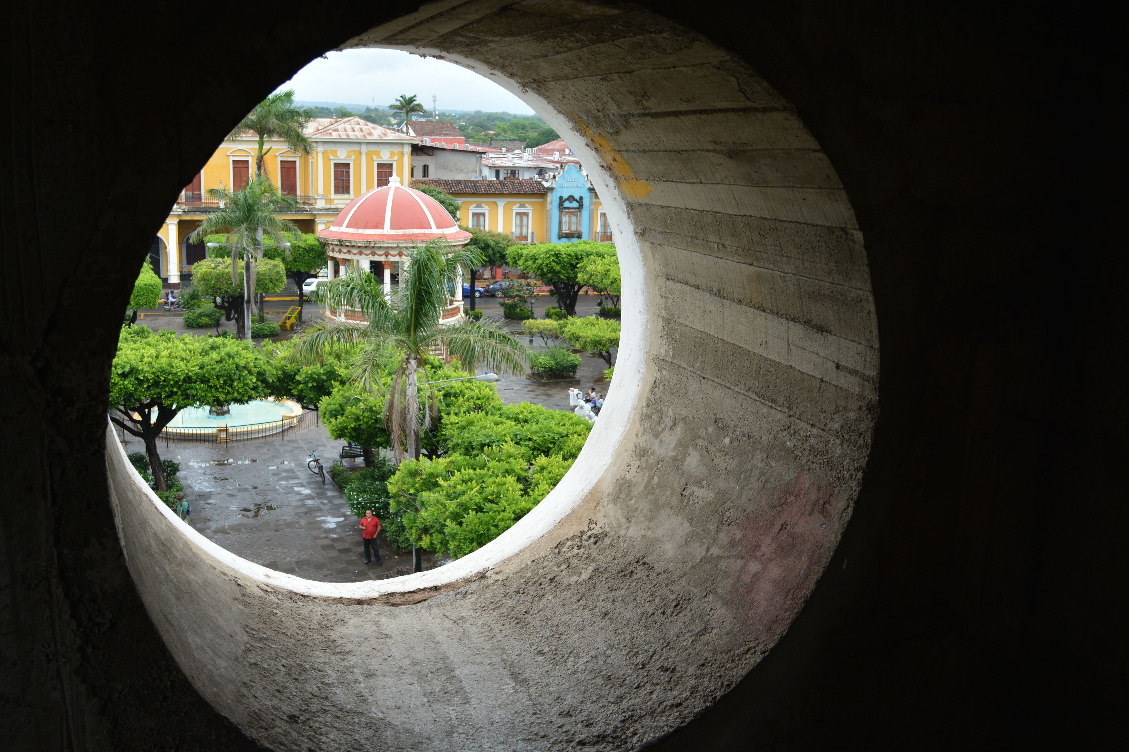 Vista a través de una ventana circular de un patio con una fuente, palmeras y edificios coloridos.